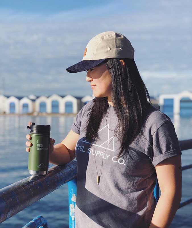 woman standing on a pier holding a stainless steel mug wearing the shades of grey adventure t-shirt.
